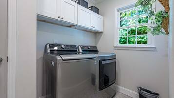 A laundry room with a front-loading washer and dryer, cabinets above, a window with a view of greenery, and a trash bin.