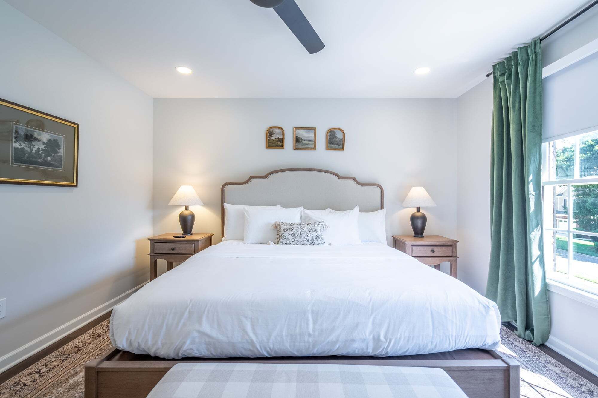 A hotel room with a large bed, two bedside tables with lamps, a ceiling fan, a window with curtains, and three framed pictures on the wall.