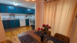 A dining area with a wooden table, brown chairs, a vase with red flowers, and a kitchen in the background.