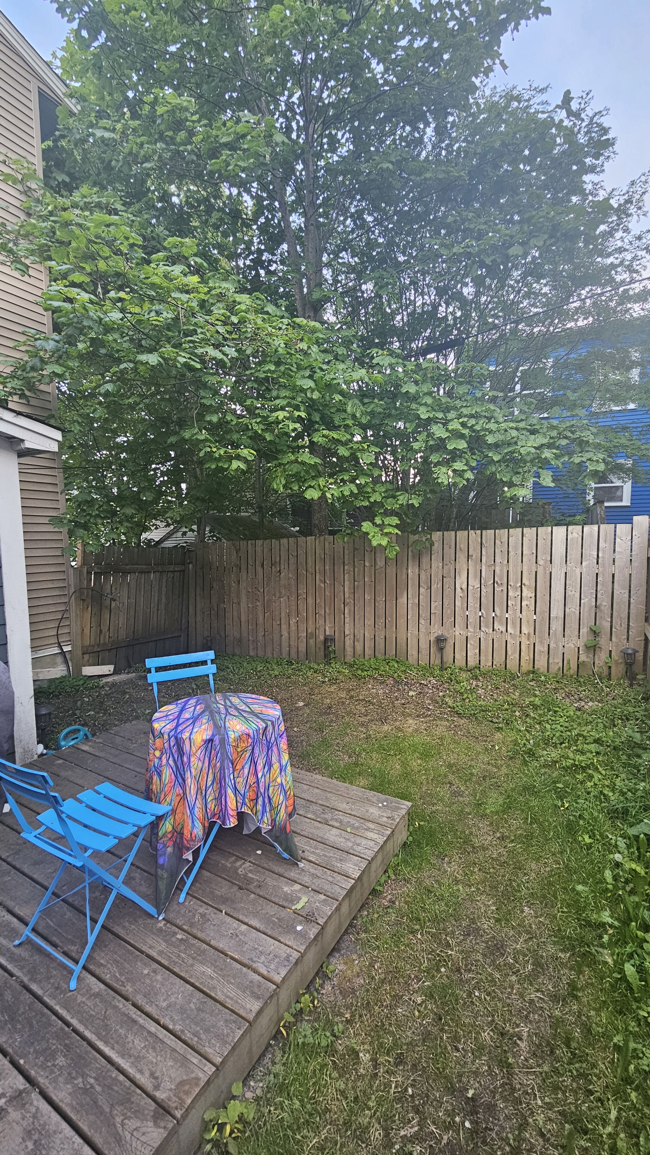 Un jardin avec une terrasse en bois, une table recouverte d’une nappe colorée et des chaises bleues.