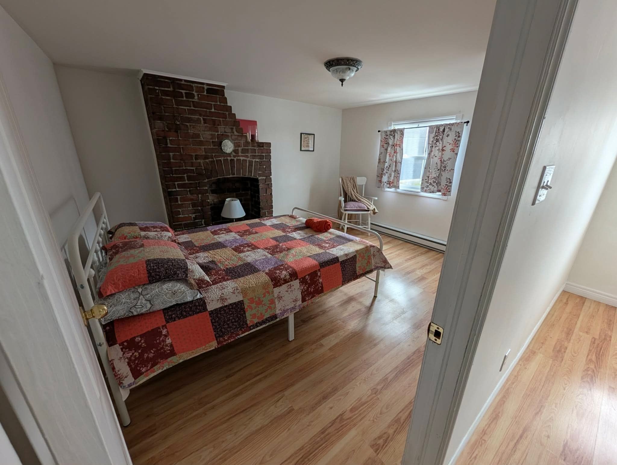 Un dormitorio con chimenea de ladrillo, una cama con colcha, una silla y un ventanal con cortinas.