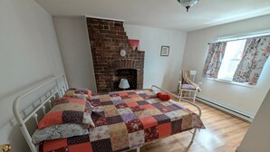 A bedroom with a brick fireplace, a bed with a colorful quilt, a chair, and a window with curtains.