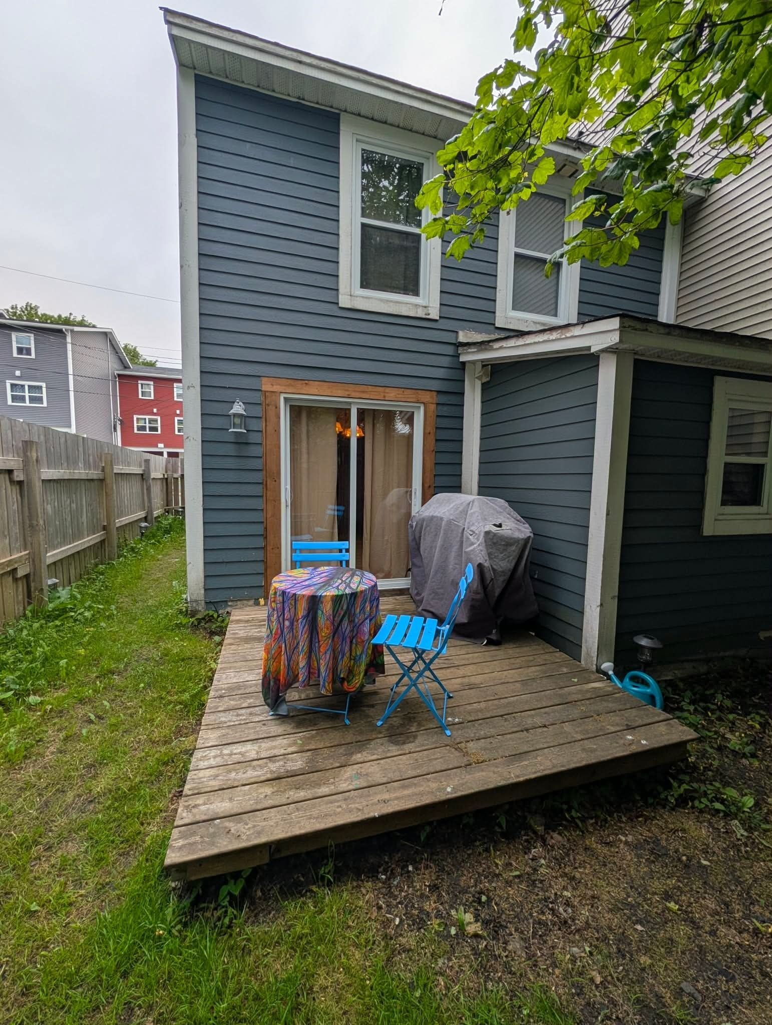 Une terrasse en bois avec une table et des chaises, un barbecue abrité et une maison à l’extérieur bleue.