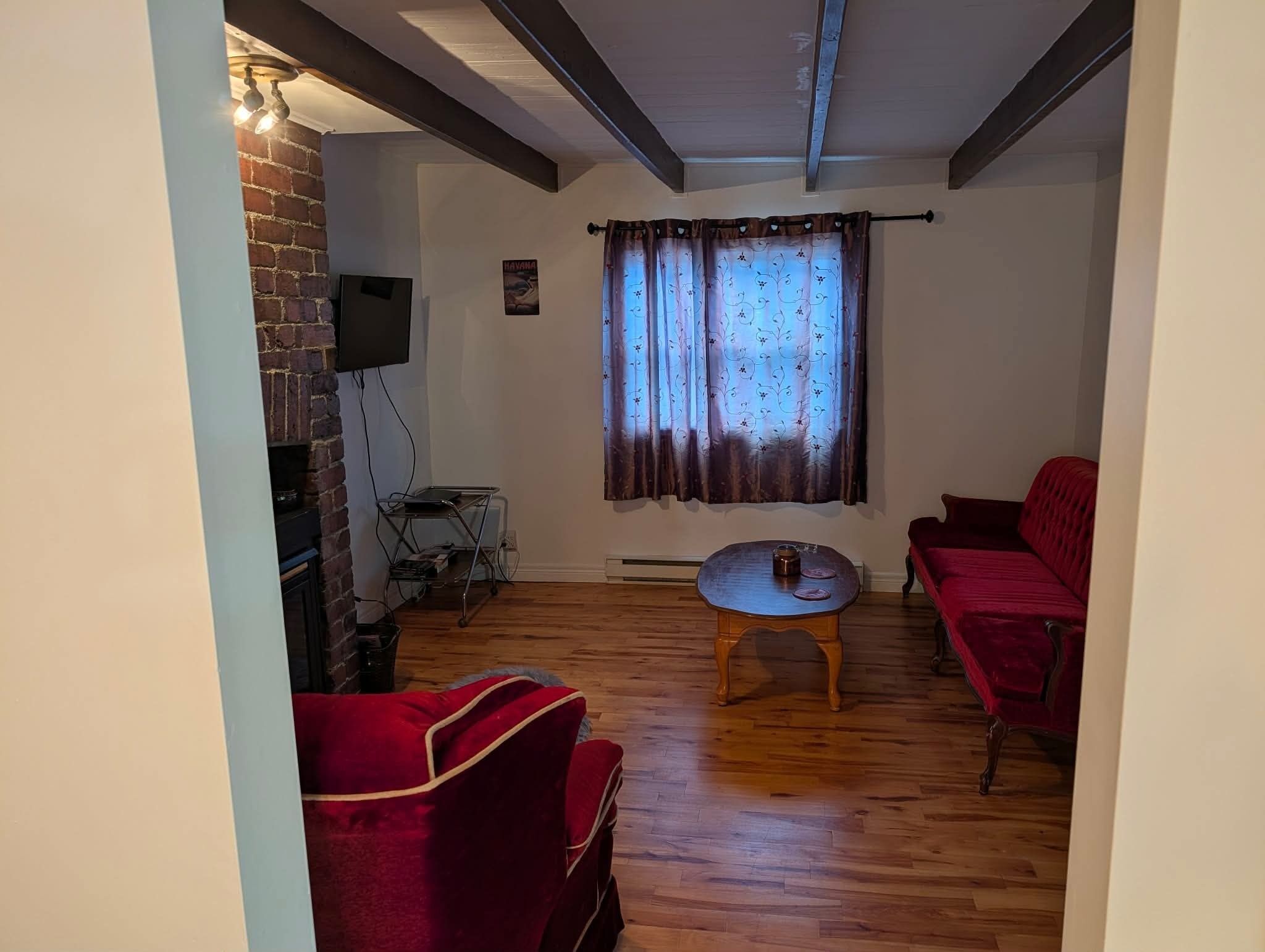 Un salon avec un mur en briques, un téléviseur à écran plat fixé au-dessus, un canapé rouge, une table basse en bois et une fenêtre ornée de rideaux à motifs.