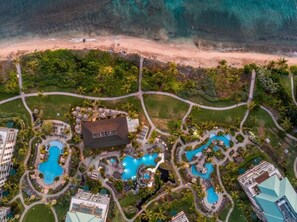 Luftaufnahme eines Resorts mit mehreren Schwimmbecken, einem großen Gebäude und einem Strand.