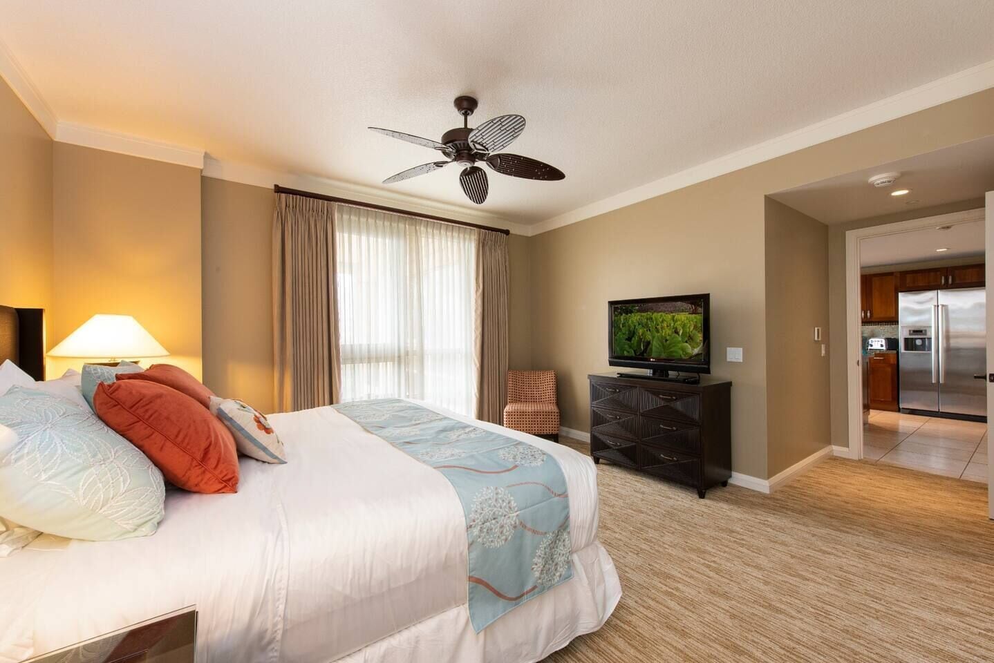 Habitación de hotel con cama, televisor en una cómoda, ventilador de techo y vista a la cocina.
