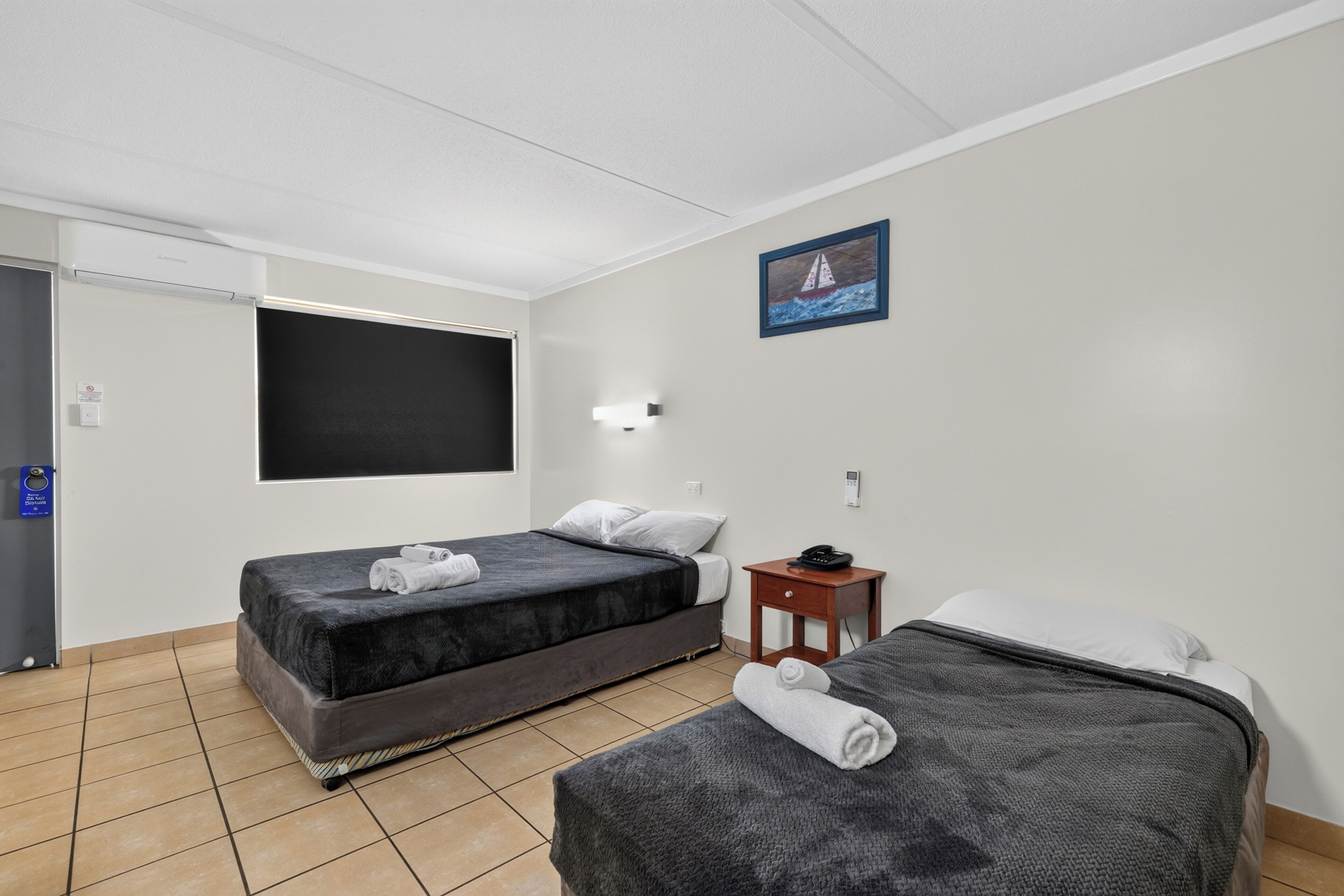 A hotel room with two beds, a flat-screen TV, a painting, and a small bedside table with a phone.