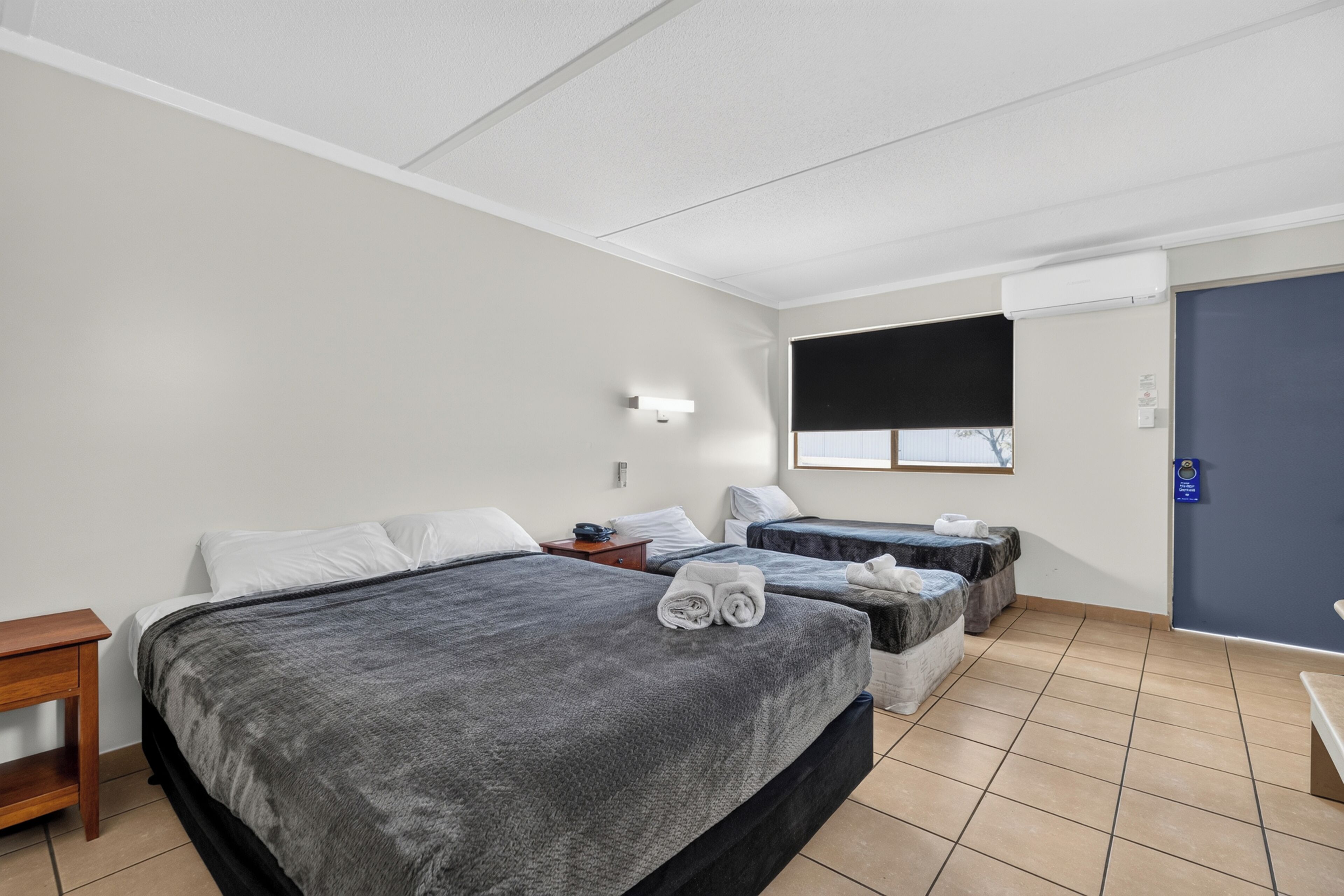 A hotel room with two beds, a desk, a window with blinds, and a door.