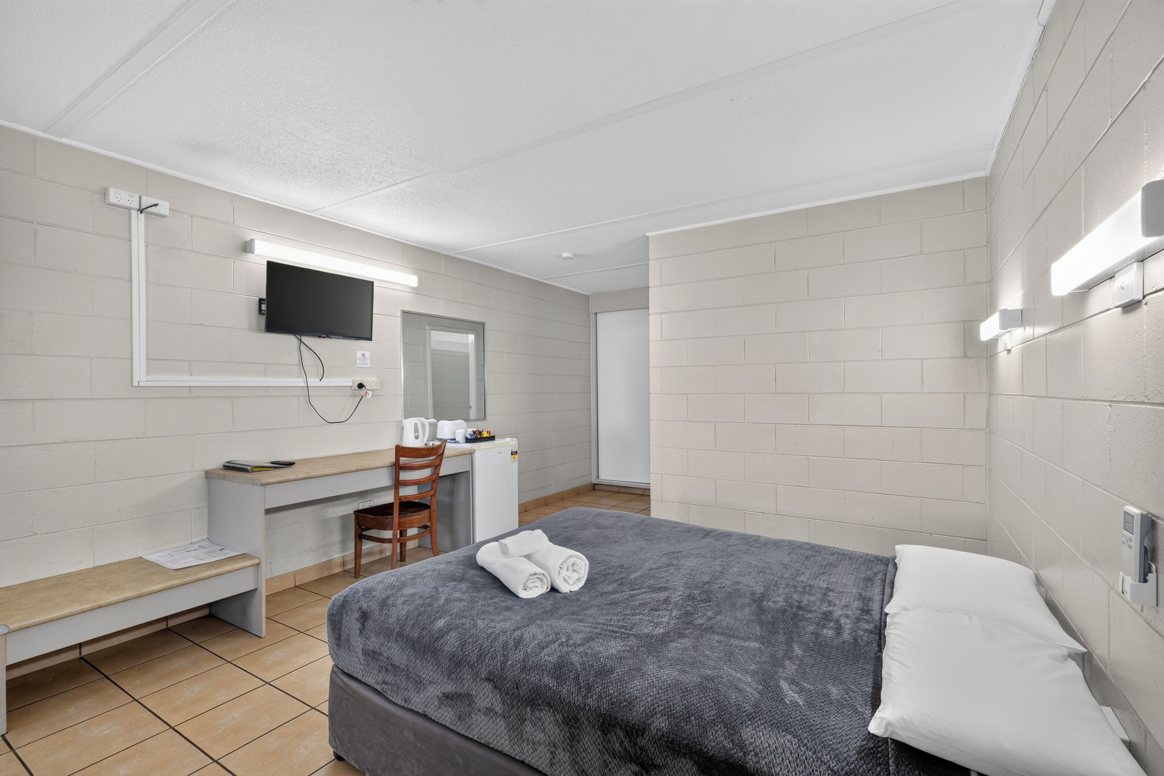 A hotel room with a bed, a desk with a chair, a television mounted on the wall, and a small kitchenette area.