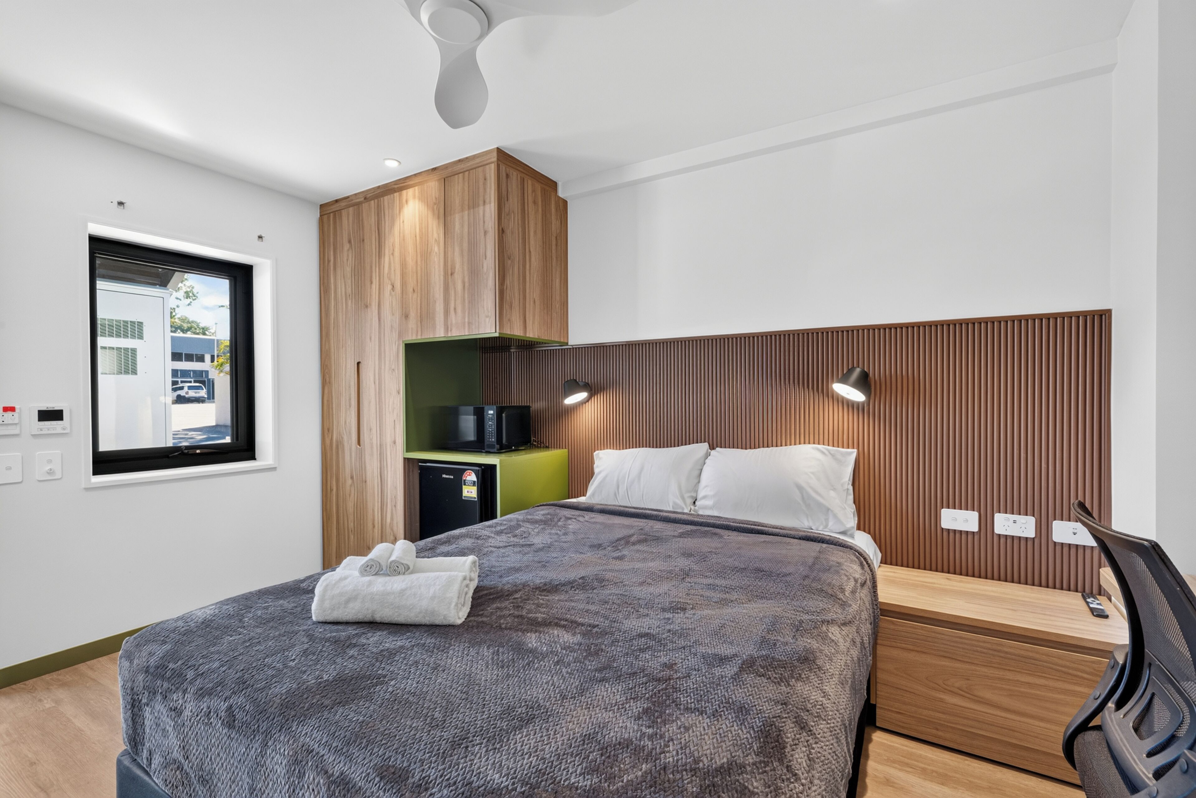 A modern hotel room with a bed, a desk, a chair, a wardrobe, and a window with a view of the outside.