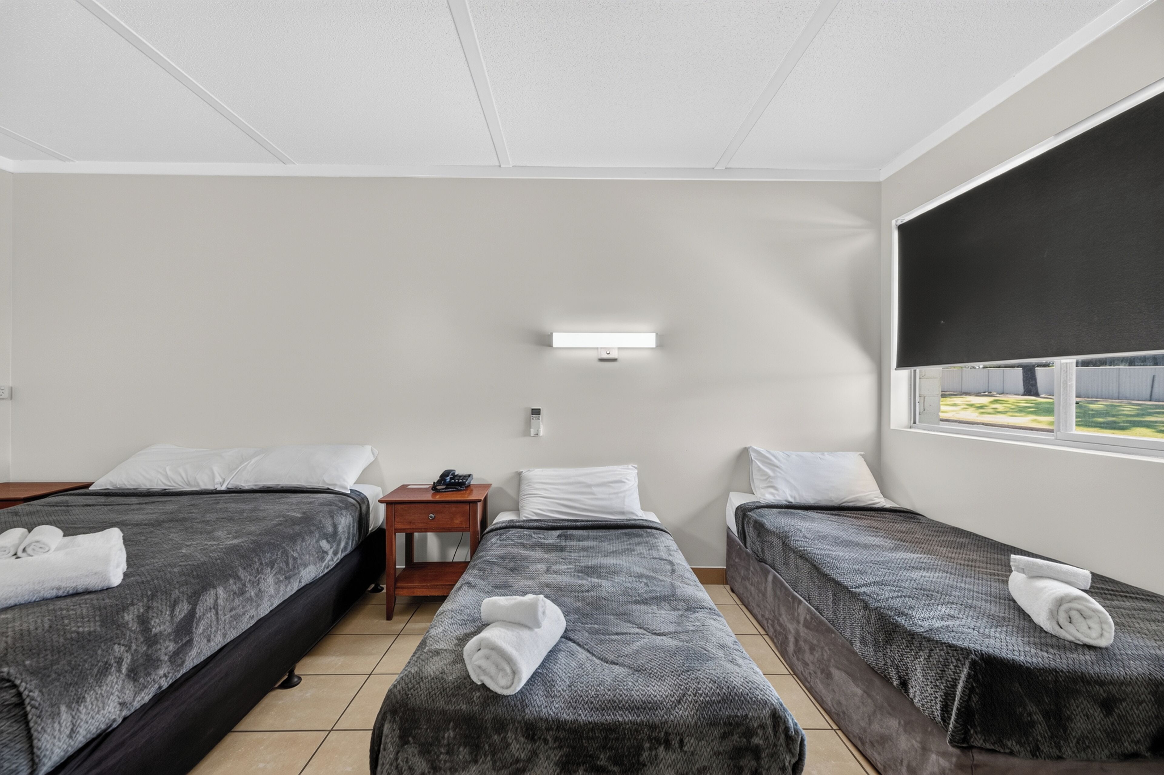 A hotel room with two beds, a small bedside table with a phone, a wall-mounted air conditioning unit, and a window with a view of greenery.