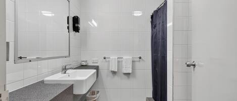Un baño con lavamanos, espejo, toallero, cortina de ducha y un cubo de basura.