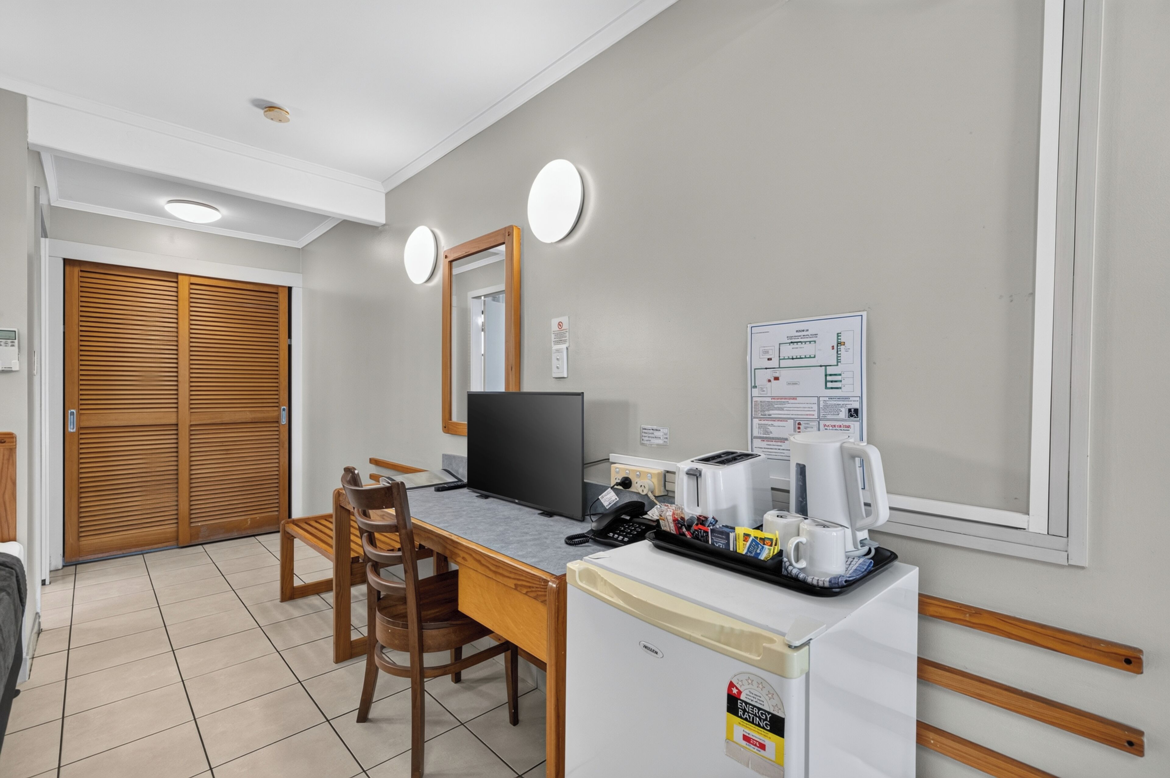 A hotel room with a wooden desk, a computer, a refrigerator, and a coffee maker.