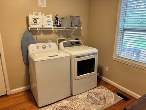 A laundry area with two front-loading washing machines and a dryer, a shelf with cleaning supplies, and a window with blinds.