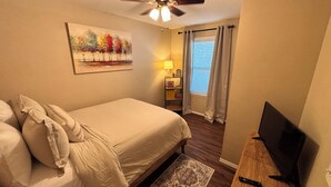 A hotel room with a bed, a television, a ceiling fan, a painting, a nightstand with a lamp, and a window with curtains. - Home Sweet Stillwater 4BR Near OSU (Stillwater)
