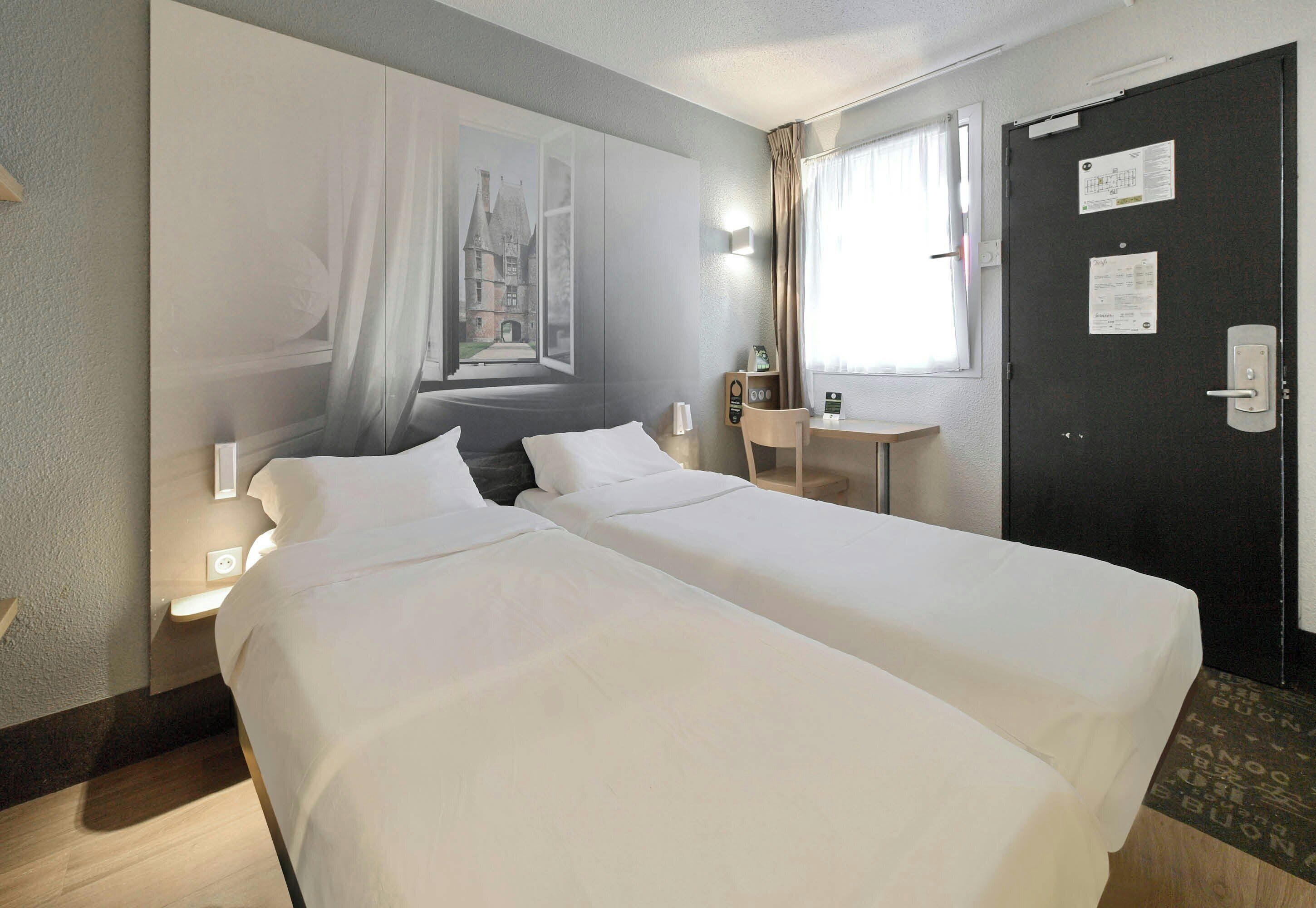 Ein Hotelzimmer mit zwei Betten, einem Fenster mit Vorhängen, einem Schreibtisch, einem Stuhl und einer Tür mit einem Hinweisschild.