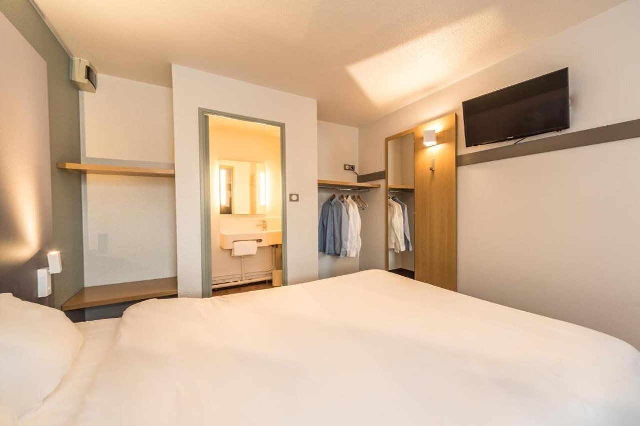Habitación de hotel con cama, baño con espejo y toalla, televisor montado en la pared y perchero.