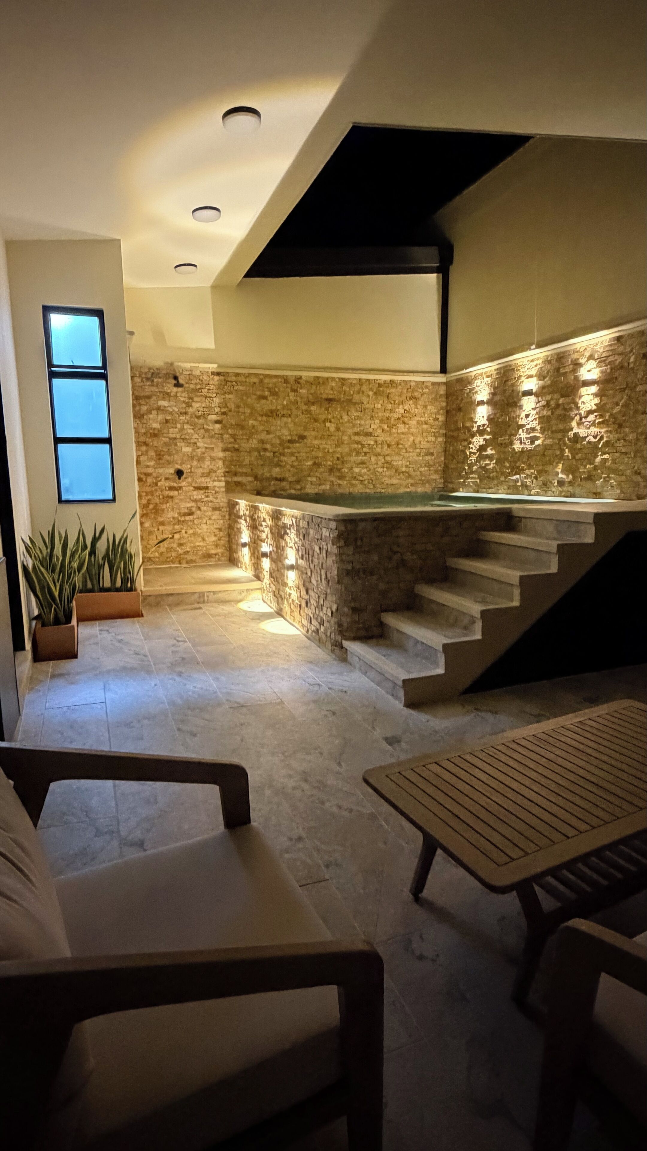 Un interior moderno con pared de piedra, una escalera y una zona de estar con un banco y una mesa.