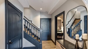 Un couloir doté d’un plancher en bois, d’une porte bleue, d’un escalier avec une rampe noire et d’un miroir qui reflète l’escalier et un mur orné d’un motif texturé.