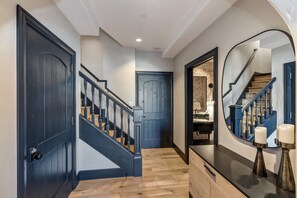 Ein Korridor mit Holzboden, einer blauen Tür, einer Treppe mit schwarzer Geländer und einem Spiegel, der eine Treppe und eine Wand mit strukturierter Musterung reflektiert.