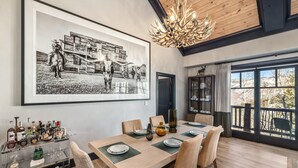 Un coin repas avec une table en bois, des chaises, un lustre, une grande photographie encadrée et un chariot à boissons avec des bouteilles.