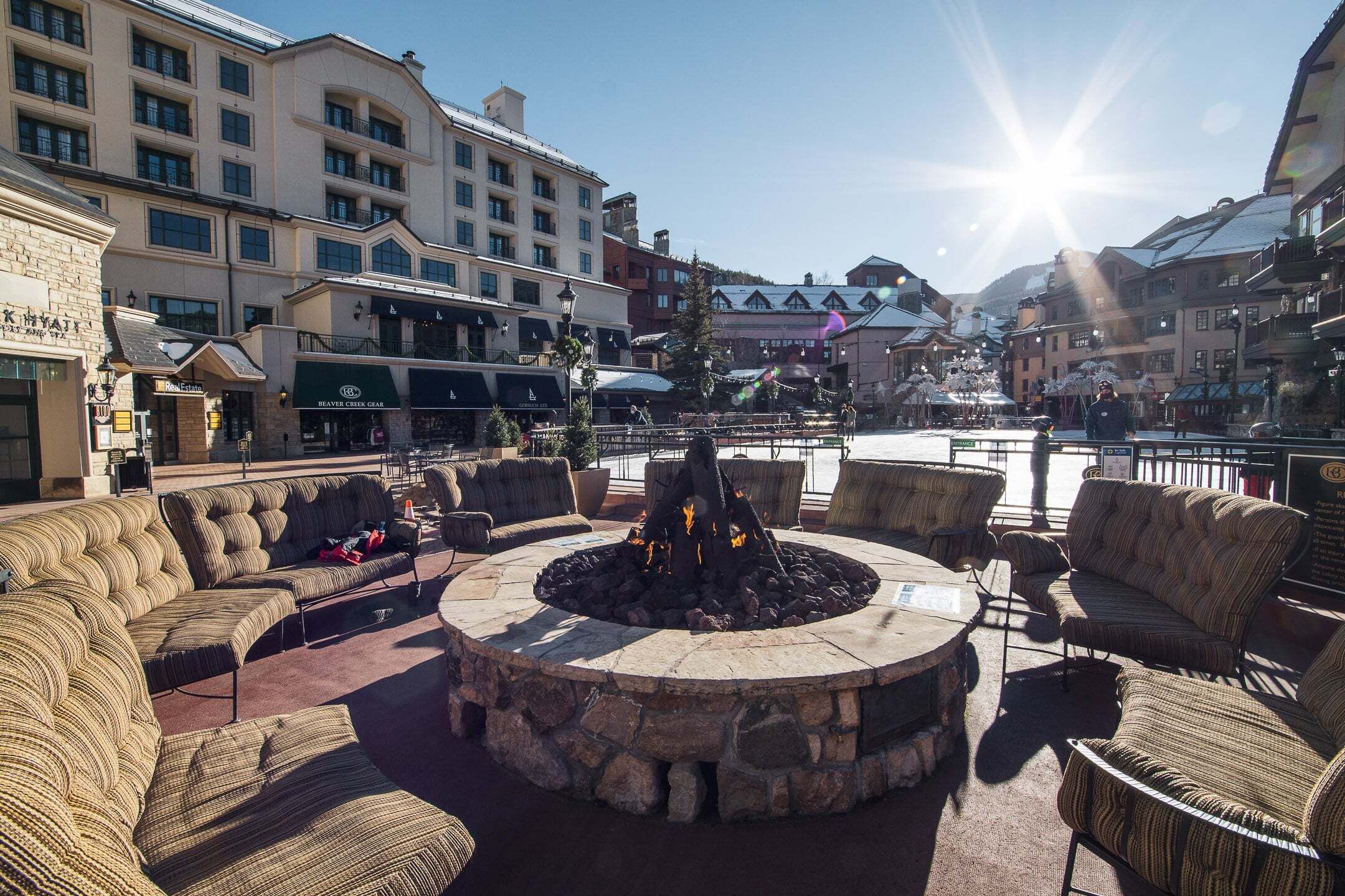 A stone fire pit surrounded by outdoor seating, a building with multiple floors, and a clear sky.