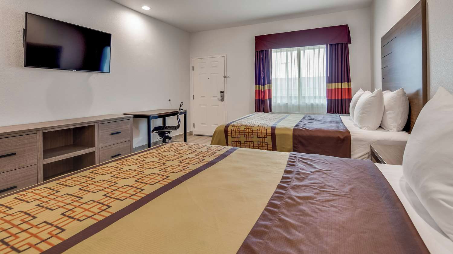 A hotel room with two beds, a desk, a television, and a window with curtains.