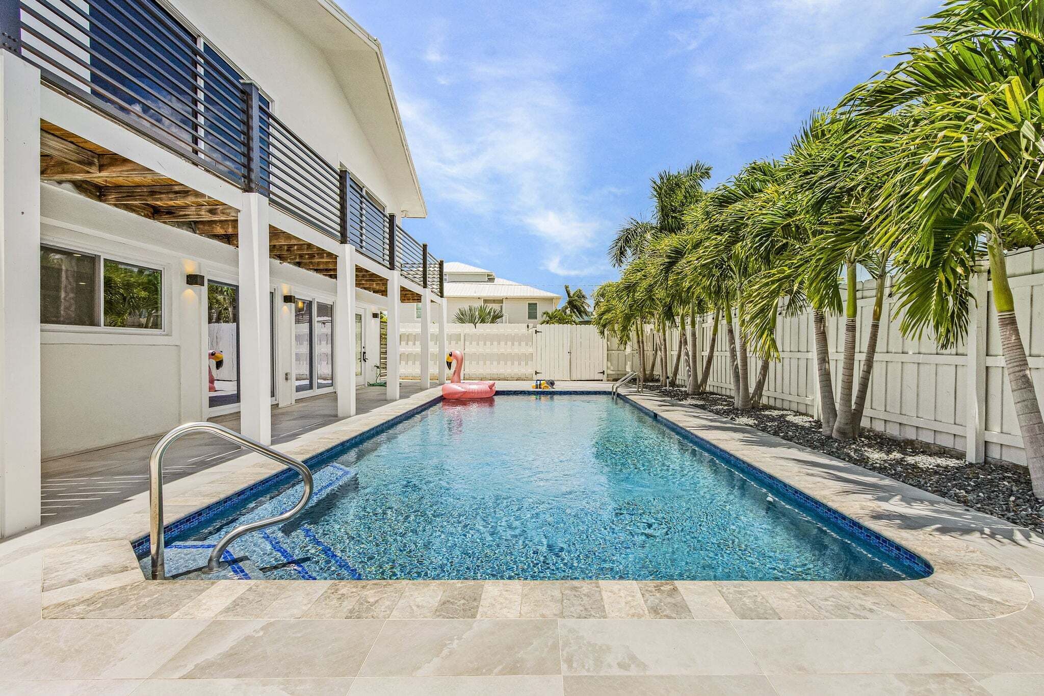 A swimming pool with a metal ladder, surrounded by a white fence and palm trees.