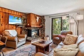 A living room with a stone fireplace, leather sofas, a television, and a large window with curtains. - Howling Wolf Winter Park Ski/Hike/Bike Condo (Winter Park)