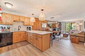 A modern kitchen with wooden cabinets, a central island, pendant lighting, and a dining area with a sofa and coffee table. - Howling Wolf Winter Park Ski/Hike/Bike Condo (Winter Park)