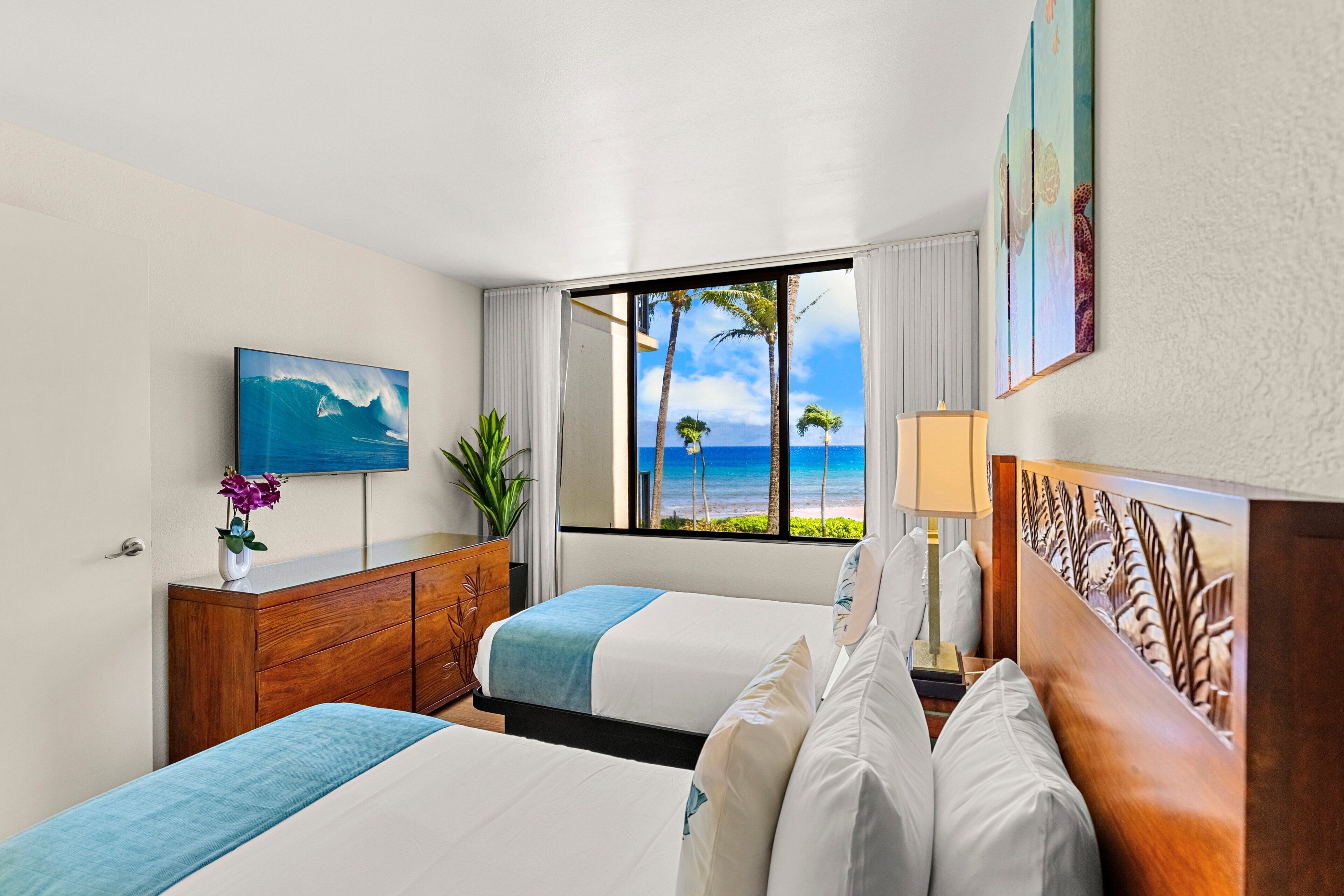 Ein Hotelzimmer mit zwei Betten, einem Fernseher, einem Fenster mit Blick auf den Strand und einer Pflanze.