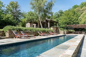 A swimming pool with lounge chairs, a stone building, and lush greenery in the background.