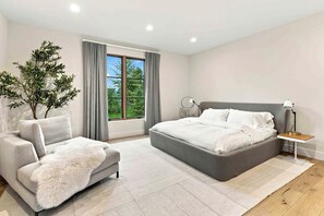 A modern bedroom with a grey bed, a grey armchair, a white fluffy rug, and a large window with curtains.