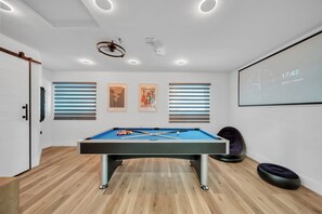 A pool table in a room with wooden flooring, a projector screen displaying a time and date, and three framed posters on the wall. - Unique 4-bedroom house with pool and gulf access in lovely Fort Myers (Fort Myers)