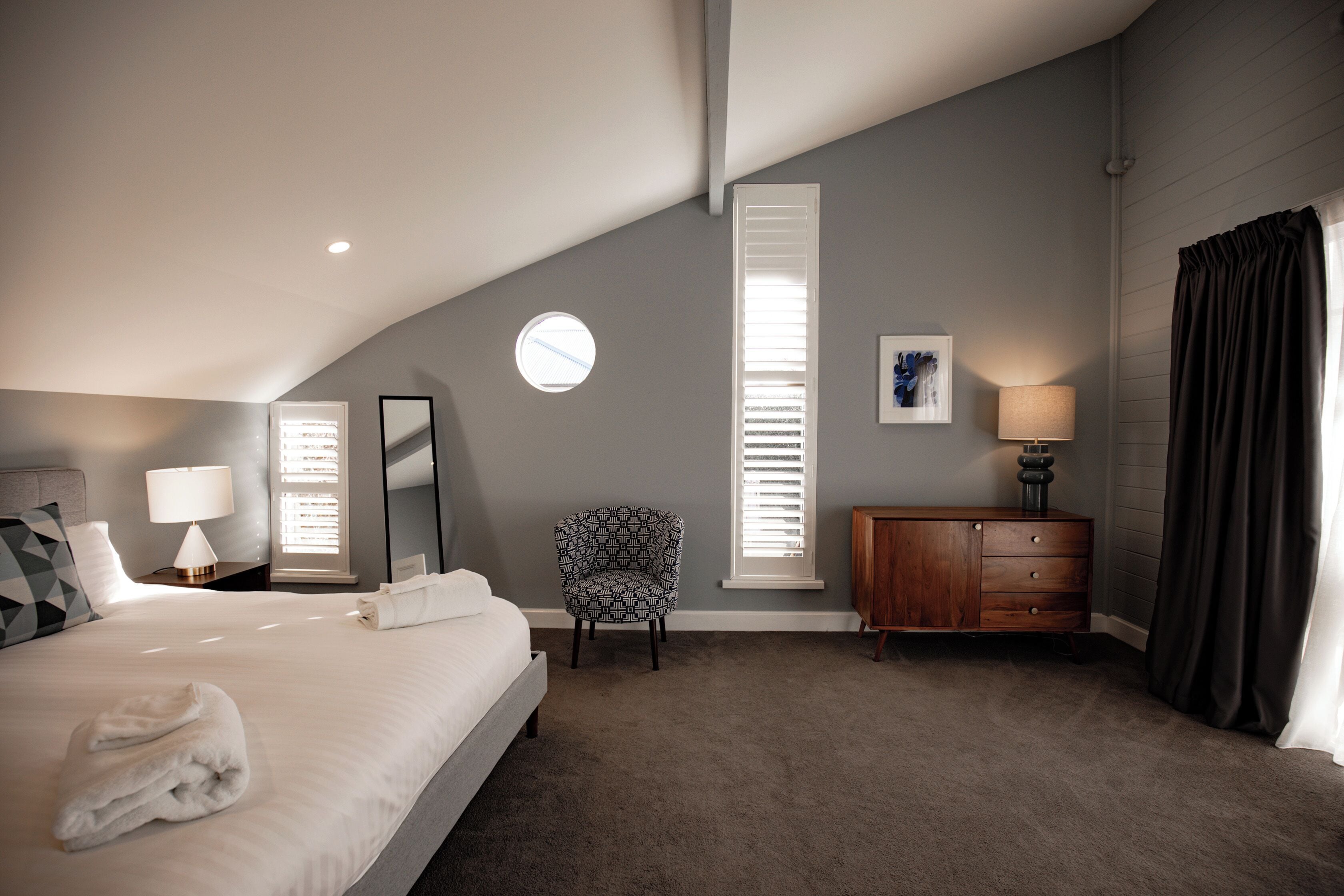 Une chambre avec un grand lit, une chaise, une commode et une fenêtre avec des stores.