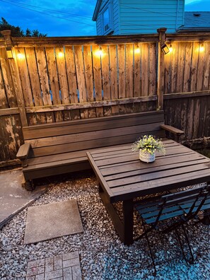A wooden deck with a bench and a table, surrounded by a wooden fence with string lights. - Cozy Detached Garden Guesthouse | Private Studio (Hamilton)