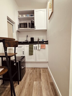 A compact kitchen with white cabinets, a microwave, and a coffee maker. There is a wooden chair and a small table with a roll of paper towels. - Cozy Detached Garden Guesthouse | Private Studio (Hamilton)