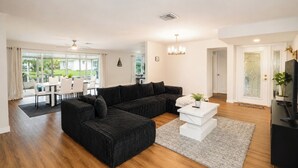 Una sala de estar moderna con un sofá seccional, un comedor con mesa y sillas blancas, y un televisor sobre un mueble negro.
