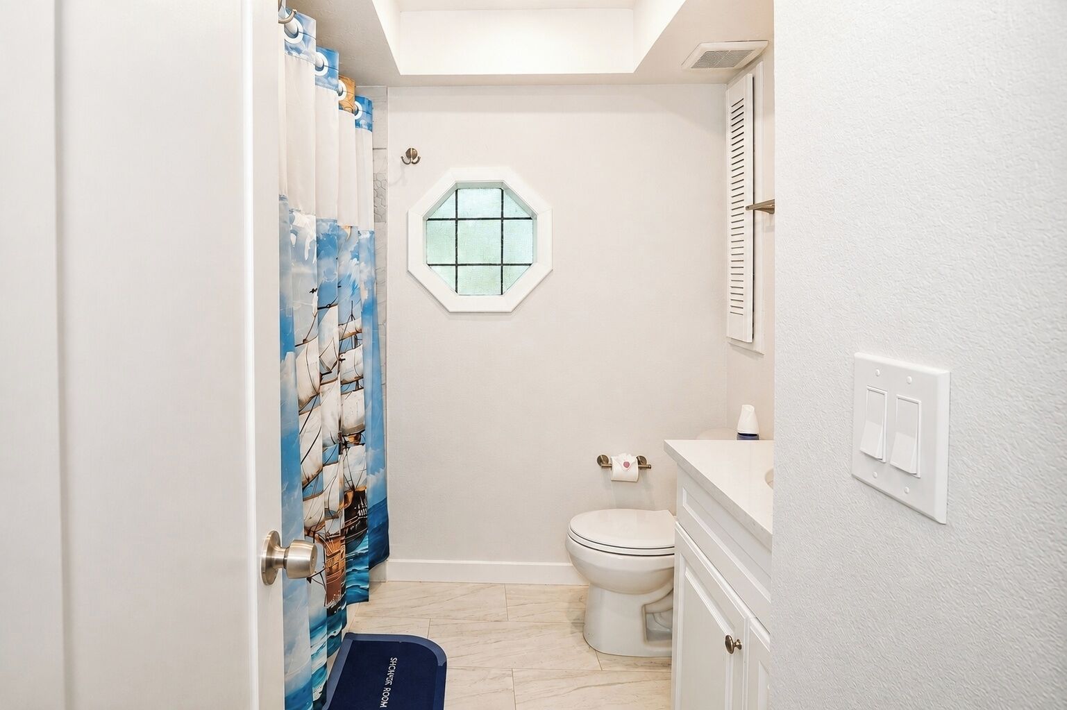 A bathroom with a toilet, a shower curtain with a nautical theme, a small window, and a towel rack.