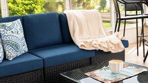 Un sofá azul con un cojín estampado, una manta blanca y una vela sobre una mesa de vidrio.