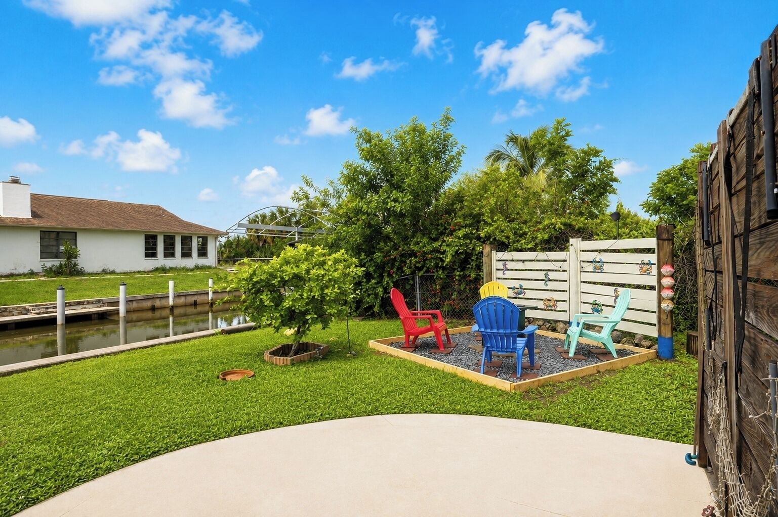A backyard with a small pond, a wooden fence, and colorful Adirondack chairs.