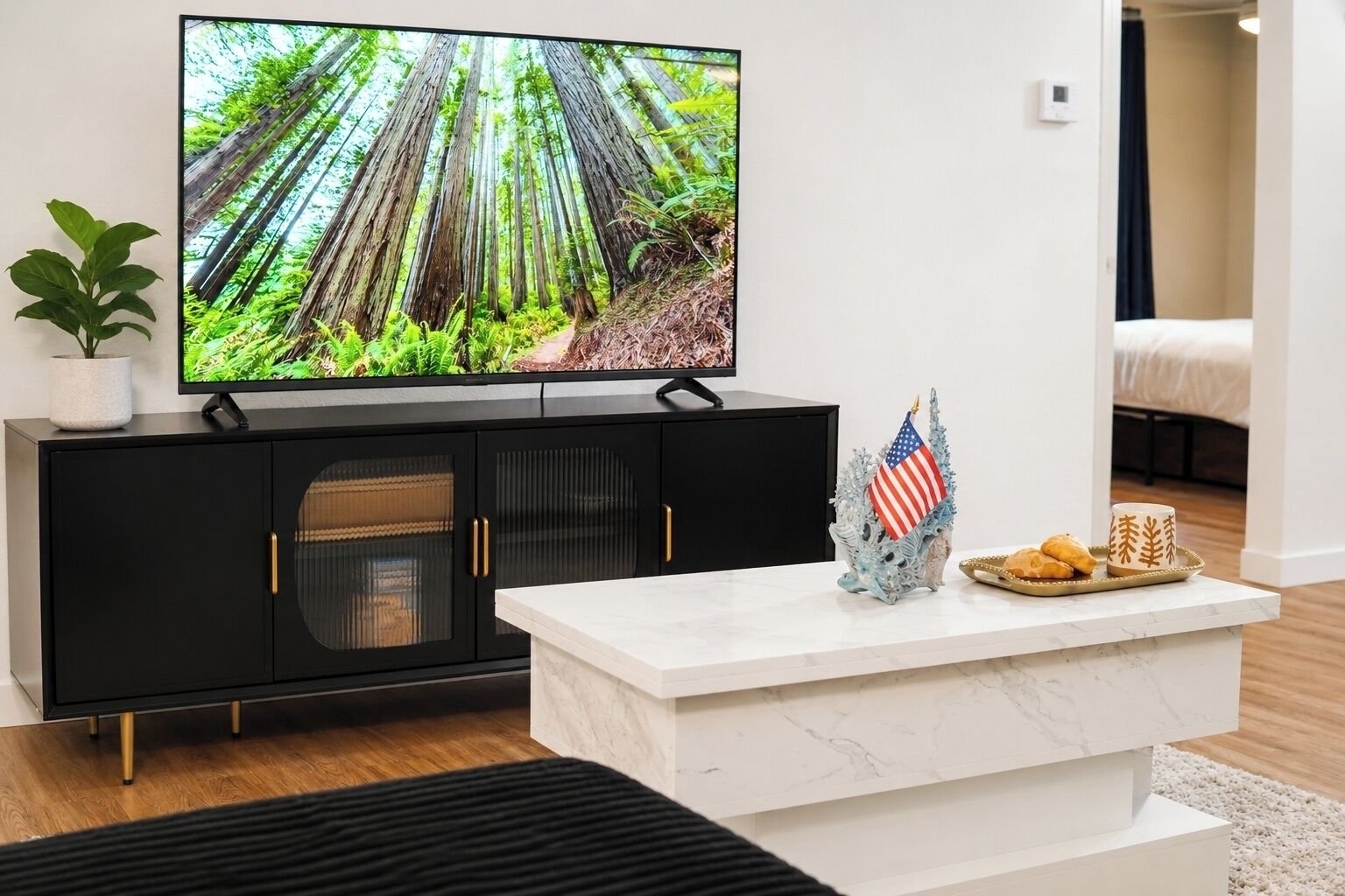 A modern living room with a flat-screen TV mounted on a black cabinet, a marble coffee table with decorative items, and a view into the adjacent bedroom.