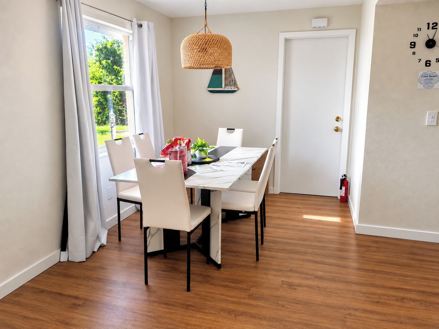 A dining area with a table, chairs, a hanging light fixture, a door, and a wall clock.