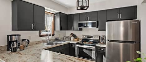 A modern kitchen with dark cabinets, stainless steel appliances, a marble countertop, and a hanging light fixture.