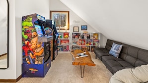 Een gezellige woonkamer met een speelhoek, inclusief een grote Marvel vs. Capcom arcadekast, een houten salontafel, een grijze bank en een boekenkast vol met gezelschapsspellen.