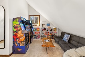 A cozy living room with a gaming area featuring a large Marvel vs. Capcom arcade machine, a wooden coffee table, a grey sofa, and a bookshelf filled with board games. - Where Fall Memories Are Made! Hiking, Campfire Nights, Games, Cozy Fireplace (Blakeslee)