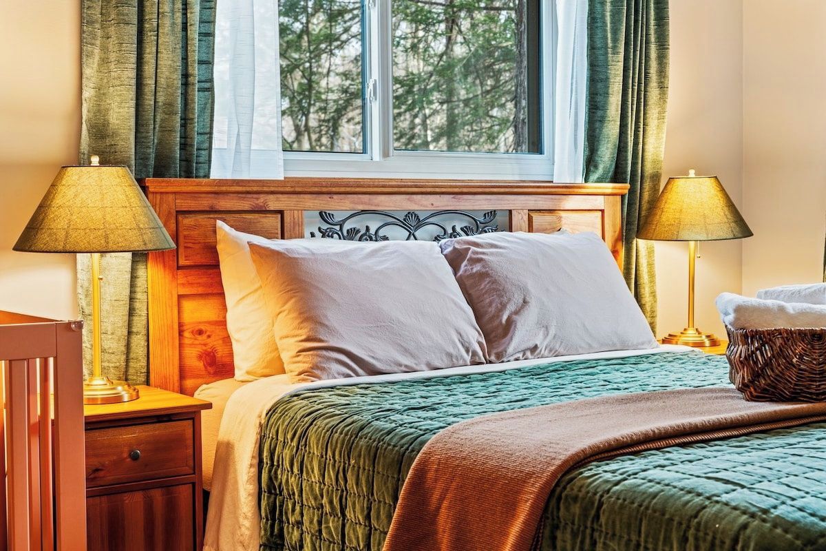 Ein Hotelzimmer mit einem Bett, zwei Nachttischlampen, einem fenster mit Vorhängen und Blick auf Bäume.