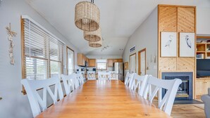 Un'ampia sala da pranzo con un lungo tavolo di legno e sedie bianche, un lampadario intrecciato e una cucina sullo sfondo.