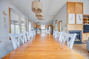 A spacious dining area with a long wooden table and white chairs, a woven light fixture, and a kitchen in the background. - 1615 - Smallfield Shores - Oceanside Corolla retreat with 4 bedrooms, ocean view, pool, hot tub. (Corolla)