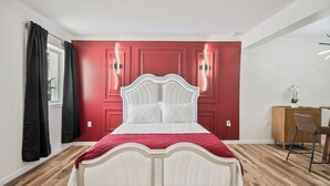 A bed with a white headboard and red bedding, a wooden floor, a desk with a chair, and a window with curtains. - Clair de Lune Studio A Charming Kokomo Stay (Kokomo)
